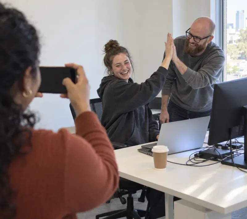 A startup co-founder holds up her phone to capture two teammates high-fiving at a laptop the moment a new feature ships.