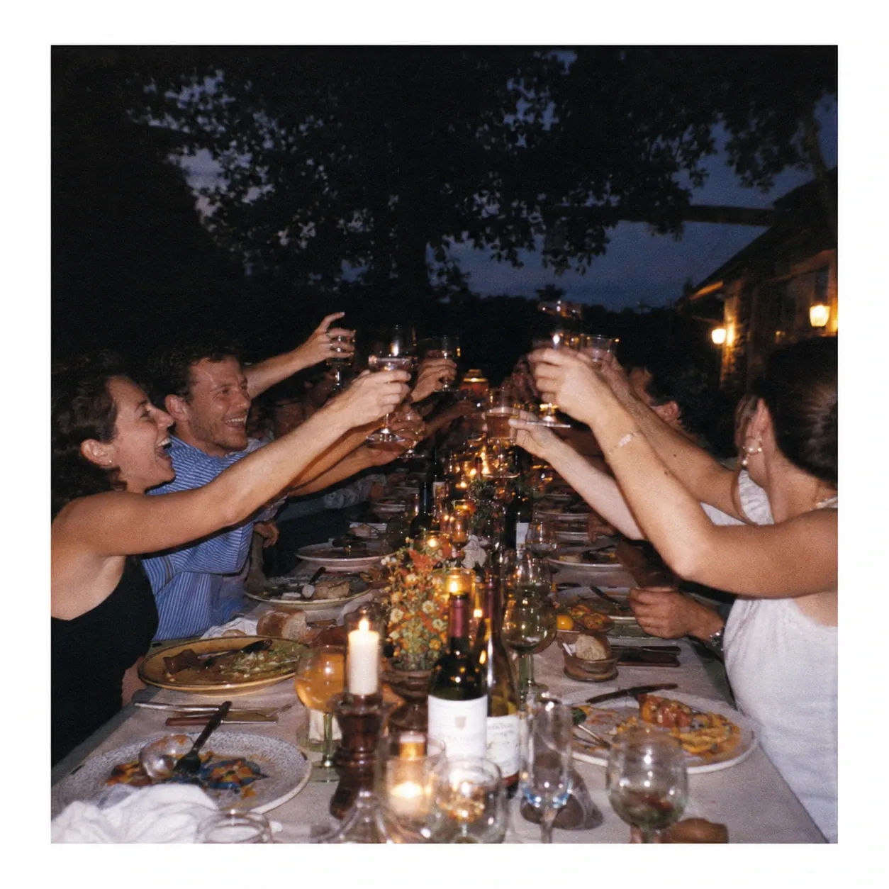 A long outdoor dinner table of friends mid-toast at dusk